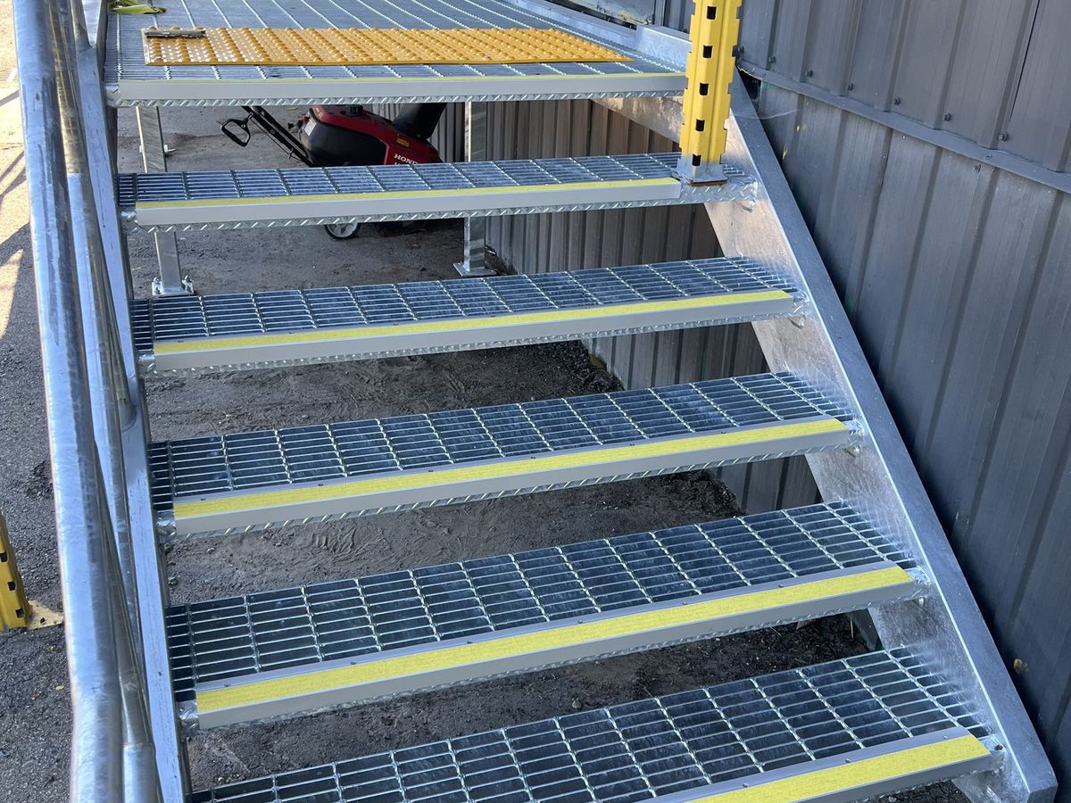 Open-grate metal stair with yellow anti-slip nosing on each tread and a tactile access tile at the top landing